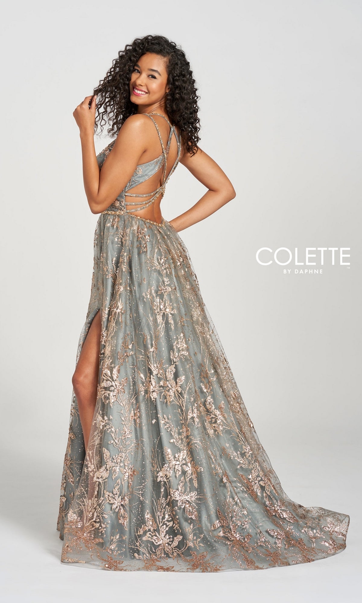 Smiling over her shoulder, a woman models the Colette CL12218 Gold and Pewter Prom Ball Gown—a sleeveless, long A-line dress with floral embroidery, strappy back, high slit, and elegant intricate detailing.